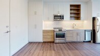 Located at 3009 W Leigh St in Richmond, Virginia, this single-floor unit showcases a clean, modern aesthetic throughout. The design emphasizes an open, airy feel with crisp white walls, recessed lighting, and consistent hardwood-style flooring that flows through the main living areas.

The kitchen is well-appointed with stainless steel appliances, including an electric range and built-in microwave. Two-toned cabinetry—combining white upper cabinets with warm wood-finish lower cabinets—adds visual depth, while a classic subway tile backsplash and open shelving provide both style and practicality.

The living area benefits from large windows offering views of the surrounding Richmond neighborhood, allowing generous natural light. A sliding barn door adds a contemporary architectural detail to one of the rooms, which also features hardwood-style flooring and a window with outdoor views. A separate carpeted room with a high clerestory window offers a quieter, more private setting.

The bathroom is finished with large-format floor tiles, subway tile surrounding the soaking tub and shower combination, and warm brass fixtures paired with a wood-toned vanity. The overall palette remains neutral and cohesive throughout, creating a calm and functional living environment.