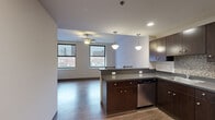 Located at 112 W Commerce St in the heart of downtown Youngstown, Ohio, this one-bedroom, one-bathroom unit offers a thoughtfully designed open-concept layout. The combined kitchen and living area forms the centerpiece of the space, featuring rich dark wood cabinetry, dark granite-style countertops, a peninsula with bar seating, and a full suite of stainless steel appliances including a refrigerator, range, microwave, and dishwasher. Pendant lighting and recessed fixtures illuminate the kitchen, while a ceiling fan keeps the living area comfortable. Hardwood-style flooring runs throughout the main living space, adding warmth and continuity. Multiple windows frame views of the surrounding downtown Youngstown skyline, filling the area with natural light. The spacious bedroom is carpeted and features dual windows that continue the urban views, creating a bright and airy atmosphere. A dedicated closet provides convenient storage. The full bathroom is finished with tile flooring, a dark wood vanity with an integrated sink, a framed mirror, and a tub-shower combination. The clean, modern aesthetic is carried consistently throughout the unit, making it a functional and comfortable urban residence in the heart of the city.