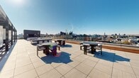Located in the vibrant Northern Liberties neighborhood of Philadelphia, this rooftop terrace offers a sweeping outdoor retreat with commanding views of the city skyline and surrounding rooftops. The large-format porcelain tile deck stretches generously across the entire rooftop level, providing ample open space for gathering, relaxing, and entertaining.

The terrace is anchored by a striking modern pergola structure, framed in sleek dark steel with warm wood slat panels that filter sunlight and create a comfortable shaded environment. Draped with flowing white curtains, the pergola shelters a central dining and lounge area complete with cushioned seating — a refined outdoor room within the open sky. Pendant globe lights add ambiance to the covered zone.

Beyond the pergola, multiple freestanding dining table sets with round tops and contoured chairs are distributed across the open deck, ideal for al fresco dining or casual gatherings. Chaise loungers invite sunbathing along the terrace perimeter, while a plush loveseat positioned near the railing offers a front-row view of the Philadelphia skyline to the south. A perimeter cable and horizontal rail safety system keeps the space secure while preserving unobstructed sightlines in every direction. The combination of open sun decks and shaded retreats makes this rooftop terrace a versatile, year-round urban amenity.