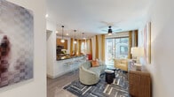 This thoughtfully designed single-floor residence combines modern aesthetics with functional living across a well-appointed open layout. The living and dining area is anchored by wide-plank wood-style flooring, a ceiling fan, and large sliding glass doors that invite abundant natural light and provide direct access to an outdoor balcony. Bold golden-yellow drapes frame the view, adding warmth and vibrancy to the space. A curved sage-green sofa and geometric area rug complete the contemporary living zone.

The kitchen opens seamlessly to the living area via a sleek breakfast bar with stylish striped stools. Warm wood cabinetry, dark countertops, a gray tile backsplash, and a full suite of stainless steel appliances — including a gas range, microwave, and refrigerator — make this kitchen as functional as it is attractive. Pendant globe lights add a refined finishing touch.

The bedroom is a bold, design-forward retreat featuring a richly upholstered yellow headboard, vibrant abstract wall art, ornate gold-framed mirrors, and dramatic patterned drapery. A ceiling fan and carpeted flooring contribute to a comfortable atmosphere.

The bathroom is clean and modern, featuring a tub-shower combination with white tile surround, wood-toned cabinetry, and dark countertops. A dedicated in-unit laundry area with a full-size washer and dryer rounds out this well-equipped residence.