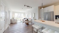 This single-floor residence is designed with a clean, contemporary aesthetic, characterized by white walls, light hardwood-style flooring, and an abundance of natural light. The open-concept layout connects the kitchen, dining, and living areas into one cohesive, flowing space.

The kitchen features a large central island with a white quartz countertop, an undermount sink, and seating for multiple bar stools. Stainless steel appliances, white cabinetry, and sleek pendant lighting complete the space. The dining area sits adjacent to the kitchen, furnished with a glass-top table and modern upholstered chairs beneath a track lighting fixture.

The living room is bright and welcoming, anchored by a large multi-pane window offering outdoor views. A glass coffee table, neutral sofa, and a teal and white area rug add texture and color to the space.

The bedroom is a calm retreat with plush carpeting, a window providing natural light, and soft neutral tones throughout. The bathroom features subway tile surrounding the tub-shower combination, a wide vanity with a white quartz countertop, and tile flooring. The entry corridor provides a smart, functional transition into the main living spaces.