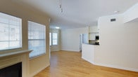 This single-floor residence offers a clean, open layout designed for comfortable everyday living. The main living area is bathed in natural light streaming through multiple windows fitted with horizontal blinds, highlighting the warm-toned hardwood flooring that runs throughout the space. Crown molding along the ceiling adds a touch of classic architectural detail, while the neutral, soft yellow walls create a welcoming and airy ambiance.

The living space flows seamlessly into a semi-open kitchen, separated by a curved breakfast bar with a dark countertop that provides both a functional workspace and a casual dining option. The kitchen is equipped with modern stainless steel appliances, including a microwave and stove, and features white cabinetry that maximizes storage while maintaining a bright, cohesive look. A flush-mount ceiling light ensures the kitchen area remains well-lit.

A hallway visible from the main living area leads to additional rooms, suggesting a practical and well-organized floor plan. The track lighting fixture above the living area provides supplemental illumination. Overall, the space presents a tidy, move-in ready interior that balances functionality with a light, open feel.