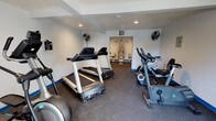 This fitness center is thoughtfully laid out across a single floor, offering a well-rounded exercise environment suited for a variety of workout routines. The main area is lined with rubber flooring and features a distinctive blue baseboard accent, providing a clean and functional aesthetic throughout. White walls and recessed lighting keep the space feeling bright and open, while wall-mounted fans ensure adequate airflow during workouts.

The primary cardio zone is generously stocked with multiple treadmills, elliptical trainers, and stationary bikes — including both upright and recumbent models — providing ample options for cardiovascular exercise. Equipment from recognized brands such as Spirit is present, indicating a focus on quality. A windowed exterior door and side windows allow natural light to filter in, creating a welcoming atmosphere.

A separate alcove off the main room serves as a dedicated strength training area, housing a multi-station cable machine, a weight bench, and additional resistance equipment. This segmented layout allows users to transition smoothly between cardio and strength training without congestion.

Exercise accessories, including stability balls, are also visible, rounding out the facility's offerings. The overall design is practical and well-organized, making this fitness center a comprehensive resource for full-body training.