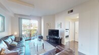 This single-floor residence offers a thoughtfully designed layout that balances comfort and functionality. The living room serves as the heart of the space, featuring rich hardwood flooring, tray ceiling details, and a large sliding glass door that opens to a private balcony with open urban views — filling the room with natural light. A leather sofa set and glass coffee table create an inviting seating area centered around a wall-mounted television.

Adjacent to the living room, the dining area accommodates a glass-top table beneath framed wall art, flowing seamlessly into the well-equipped kitchen. The kitchen is outfitted with dark cabinetry contrasted by light countertops, stainless steel appliances including a range, microwave, dishwasher, and refrigerator, all set on hardwood flooring with recessed lighting overhead.

The bedroom provides a serene retreat with plush carpeting, a large upholstered headboard, ample natural light from a bright window, and a dedicated workspace. Two bathrooms serve the residence: the primary bathroom features a wide vanity with backlit mirrors and a glass-enclosed shower, while the second bathroom includes a full tub-shower combo with classic finishes and hardwood flooring. Clean white walls and consistent design choices throughout lend the space a cohesive, modern ambiance.
