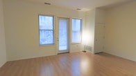 This single-floor space offers a clean, modern interior designed with simplicity and functionality in mind. The main living area is anchored by smooth light hardwood flooring that reflects natural light and lends a warm, airy feel throughout. Two generously sized windows flank a glass-paneled door, together creating an exceptional amount of natural light while providing views of the neighborhood outside. The windows are fitted with adjustable blinds for privacy and light control. The walls are finished in a neutral, light tone that complements the flooring and enhances the sense of openness. Ceiling-mounted ventilation registers indicate a central HVAC system, ensuring climate comfort year-round. A floor vent near the exterior wall further supports efficient heating and cooling. The layout flows openly, with a visible hallway leading to additional areas of the unit, including what appears to be a closet or storage space. The overall ambiance is minimal and well-lit, making the space highly adaptable for a variety of uses. The combination of natural light, practical finishes, and a straightforward layout gives this space a welcoming and versatile character.