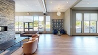This single-floor commercial space offers a generously sized, open-concept layout designed to function as a welcoming lobby and multi-purpose lounge area. The interior is defined by warm hardwood flooring that runs throughout, complemented by neutral-toned walls in muted blues and greiges that lend a contemporary, professional atmosphere.

A striking stacked-stone feature wall serves as a central focal point, housing a linear gas fireplace that adds warmth and visual interest to the space. Large floor-to-ceiling windows along the exterior walls flood the interior with abundant natural light while providing views of the surrounding outdoor environment. The varied ceiling design incorporates recessed lighting, exposed metallic ductwork, and elegant cluster-style pendant chandeliers that balance industrial and refined aesthetics.

The space is organized into distinct functional zones, including a lounge area with plush sofas and accent chairs, a dining or meeting area with a wooden communal table and metal chairs, and a more intimate seating nook with club chairs. An Interac ATM is conveniently located near the staircase. Wall-mounted artwork and a grid-style display panel add curated decorative touches, reinforcing the polished, hospitality-forward character of the space.