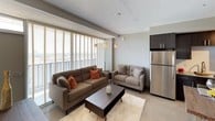 This well-appointed single-floor residence showcases a thoughtfully designed layout with modern finishes throughout. The open living area is bathed in natural light streaming through expansive floor-to-ceiling windows fitted with horizontal blinds, offering sweeping elevated views of the surrounding cityscape. The seating area is anchored by a comfortable sectional sofa and loveseat arrangement, creating an inviting space ideal for relaxing or entertaining.

The kitchen is designed for both functionality and style, featuring dark espresso cabinetry, granite countertops, a stainless steel undermount sink, and a full suite of stainless steel appliances including a range, microwave, and refrigerator. Track lighting overhead enhances the galley-style layout, which flows seamlessly toward the entryway.

The residence includes two bedrooms, each finished in soft neutral tones. The primary bedroom benefits from large windows with outdoor views, while the secondary bedroom features unique angled ceiling details and warm accent lighting. Both rooms offer a calm, restful atmosphere.

The bathroom is clean and contemporary, featuring a large walk-in shower enclosed by a frameless sliding glass door and clad in white large-format tile. A dark wood vanity with a granite countertop and a polished concrete floor complete the sophisticated look. An adjacent walk-in closet adds practical storage to the space.