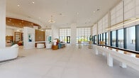 Located in Saint Petersburg, Florida, this expansive single-floor amenity space offers a sophisticated and airy environment designed for both relaxation and recreation. The interior is defined by soaring double-height ceilings, large-format white tile flooring, and dramatic floor-to-ceiling windows fitted with layered roller shades that bathe the space in abundant natural light while framing views of the lush outdoor grounds and pool area beyond.

The open-plan layout is anchored by a central lounge area featuring a curated mix of contemporary seating — including rounded white chairs, teal accent sofas, and warm wood side tables — arranged atop textured area rugs. A warm wood-paneled feature wall with a graceful arched detail and built-in shelving provides an elegant focal point, complemented by colorful abstract artwork throughout.

The game room area is a standout highlight, equipped with a sleek white pool table beneath a gold oval pendant light and a full-size shuffleboard table, alongside arcade games for added entertainment. Bar-height seating with gold accents lines one section of the space, ideal for social gatherings. Multiple glass door entries connect seamlessly to the outdoor terrace and pool deck, reinforcing the bright, resort-style ambiance that defines this well-appointed community amenity space.