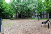 This outdoor dog park is a well-appointed amenity space designed to provide pets and their owners with a safe and comfortable environment. The area is fully enclosed by a sturdy black chain-link fence, complete with a gated entry point that ensures pets can roam freely without the risk of escape. The ground is covered with a generous layer of wood chip mulch, offering a soft, low-maintenance surface suitable for active play. A grassy buffer runs along the perimeter of the fenced enclosure, adding a touch of greenery to the space. A metal mesh bench is conveniently positioned within the park, giving owners a comfortable spot to sit and supervise their pets during visits. Mature, densely canopied trees surround the area, providing natural shade and a tranquil, wooded atmosphere. The backdrop features multi-story residential buildings with balconies and staircases, integrating the dog park seamlessly into the broader community setting. The combination of practical fencing, a soft ground cover, seating, and abundant greenery makes this dog park a functional and inviting destination for residents and their four-legged companions.