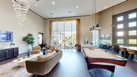 Located at 18235 Bulverde Rd in San Antonio, TX, this single-floor community clubhouse offers a generously sized, open-concept gathering space designed with modern sophistication. Rich hardwood-style flooring runs throughout, anchoring a cohesive aesthetic that blends comfort with contemporary style.

The main lounge area features plush sofas, accent chairs, and a central billiards table, creating a versatile space for both relaxed socializing and entertainment. Adjacent to the lounge, a long counter-height bar lined with leather barstools sits alongside floor-to-ceiling glass partitions, offering a sleek co-working or social perch with views into the surrounding space.

Natural light floods the interior through expansive floor-to-ceiling windows, framing outdoor views of lush landscaping and the surrounding community. Soaring ceiling heights amplify the sense of openness throughout. A dedicated meeting or office nook features a round conference table, task chairs, and a tiered grid of clerestory windows that draw in additional daylight.

Sophisticated lighting elements—including sculptural ring chandeliers and pendant fixtures—add a refined, upscale touch. A refreshment station and flat-screen display area further enhance the space's functionality. The overall ambiance is polished, welcoming, and well-suited for community gatherings, remote work, or casual lounging.