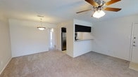 This single-story space features an open-concept layout that seamlessly connects the living area to the kitchen. The living room is finished with wall-to-wall neutral-toned carpeting, creating a comfortable and cohesive feel. A ceiling fan with an integrated light fixture is centrally positioned overhead, providing both airflow and ambient lighting to the space.

The kitchen is defined by a half-wall peninsula with a light stone countertop, offering a natural dividing point between the cooking and living areas. Dark upper cabinetry contrasts with the white walls, while stainless steel appliances, including a refrigerator and microwave, add a modern touch. The kitchen's compact, efficient design maximizes functionality within the open floor plan.

A pendant light fixture hangs near what appears to be a hallway or entryway corridor, leading further into the unit. The walls throughout are painted in a clean, bright white, enhancing the sense of openness. Neutral tones dominate the overall palette, contributing to a simple, versatile ambiance that allows for a variety of furnishing and décor styles.