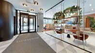 Located at 2812 Rue Ontario E in Montréal, this single-floor amenity space offers a sophisticated and thoughtfully designed environment spanning a generous footprint. The space is anchored by a striking central lobby featuring large-format light grey tile flooring, exposed concrete columns, and floor-to-ceiling windows that flood the interior with natural light and provide views of the surrounding streetscape. A glass-partitioned entry displays a curated open shelving unit adorned with lush trailing plants, books, and decorative objects, establishing an immediate sense of warmth and refinement upon arrival. The main lounge area is defined by a circular communal table with a built-in planter at its center, surrounded by pendant lighting with a warm glow. Terracotta-toned seating pods and accent chairs are arranged throughout, complemented by rich orange carpeted zones that contrast elegantly with the neutral tile. Warm wood paneling and burnt-orange wall accents add depth to the overall palette. A dedicated private office features acoustic wall panels, a clean-lined workstation, and access to natural light, while a compact focused-work room provides an intimate, distraction-free setting. Together, the areas offer a seamless blend of collaborative and private functionality within a vibrant, biophilic-inspired design.