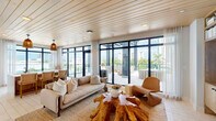 Located in the heart of Fort Myers, FL, this thoughtfully designed single-floor space combines contemporary architecture with warm, organic materials to create an inviting and sophisticated atmosphere. The interior living area features a striking wood-planked ceiling that adds warmth and texture, complemented by expansive floor-to-ceiling black-framed windows and glass doors that flood the space with natural light and provide seamless views of the outdoor terrace and city skyline. Natural fiber rugs, a live-edge teak coffee table, and a mix of neutral-toned and woven furnishings reinforce the space's organic aesthetic. A built-in shelving unit with integrated media display and a dining area with cane-back chairs round out the interior, offering both function and style. A standout feature is the dedicated outdoor living area, sheltered beneath a bold black pergola structure and adorned with string lights. This terrace is furnished with a generous modular sectional sofa and rustic teak stump side tables, all oriented to take full advantage of the sweeping views of Fort Myers' skyline and lush tropical palm canopy. An additional area showcases a dramatic large-format natural stone accent wall, adding an earthy, textural focal point. Together, these spaces deliver a cohesive blend of coastal-modern design and natural elegance.