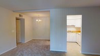This single-floor space features an open-concept layout that seamlessly connects the living, dining, and kitchen areas, creating a cohesive and functional environment. The dining area is defined by a elegant chandelier that serves as a focal point, casting warm light over the carpeted floor below. Neutral-toned walls throughout lend a clean, versatile backdrop suitable for a variety of interior styles.

A structural column partially separates the living and dining zones while maintaining the open feel of the space. The kitchen is situated adjacent to the main living area and is equipped with white cabinetry, white appliances including a refrigerator, and a sink set into a countertop with a light wood finish. The white cabinetry features decorative molding along the upper cabinets, adding a classic touch to the kitchen design.

The flooring transitions from carpet in the living and dining areas to a hard surface in the kitchen, clearly delineating each functional zone. Recessed soffits frame the dining area, adding subtle architectural definition. Overall, the space presents a straightforward, practical layout with traditional design elements throughout.