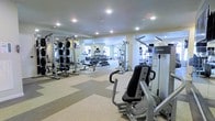 This fitness center offers a comprehensive workout environment designed to accommodate a wide range of exercise needs. The space is outfitted with a solid selection of strength-training equipment, including multi-station cable machines, a free weight rack with a full dumbbell set, and dedicated weight benches — providing ample options for resistance training. Cardio equipment, including treadmills, is positioned along the perimeter, visible in the mirrored walls that line the room and serve to visually expand the space while allowing users to monitor their form. The flooring consists of a durable, light-toned rubber or vinyl surface that provides both traction and comfort underfoot. Recessed ceiling lights are evenly distributed throughout, delivering consistent, bright illumination across the entire floor area. The neutral-toned walls and clean layout contribute to an organized, functional atmosphere. The mirrors not only enhance the sense of space but also reflect natural light from windows visible in the background, adding a welcoming brightness to the environment. Overall, the fitness center is clean, well-maintained, and thoughtfully arranged to support an effective and comfortable workout experience.