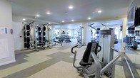 This fitness center offers a comprehensive workout environment designed to accommodate a wide range of exercise needs. The space is outfitted with a solid selection of strength-training equipment, including multi-station cable machines, a free weight rack with a full dumbbell set, and dedicated weight benches — providing ample options for resistance training. Cardio equipment, including treadmills, is positioned along the perimeter, visible in the mirrored walls that line the room and serve to visually expand the space while allowing users to monitor their form. The flooring consists of a durable, light-toned rubber or vinyl surface that provides both traction and comfort underfoot. Recessed ceiling lights are evenly distributed throughout, delivering consistent, bright illumination across the entire floor area. The neutral-toned walls and clean layout contribute to an organized, functional atmosphere. The mirrors not only enhance the sense of space but also reflect natural light from windows visible in the background, adding a welcoming brightness to the environment. Overall, the fitness center is clean, well-maintained, and thoughtfully arranged to support an effective and comfortable workout experience.