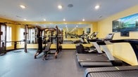 This fitness center is a comprehensively outfitted exercise facility designed to support a wide range of workout routines. The space is immediately distinguished by its bold yellow walls, which lend an energetic ambiance, complemented by a speckled rubber floor that provides durability and slip resistance underfoot.

The cardio zone features multiple treadmills and a stationary bike, each equipped with individual display screens for an engaging workout experience. A wall-mounted flat-screen television is also present, adding entertainment options for users. Natural light filters in through several windows fitted with blinds, contributing to a bright and welcoming environment.

The strength training area is well-stocked, offering a multi-station cable machine, an adjustable weight bench, and a fully loaded dumbbell rack with kettlebells and medicine balls stored on dedicated shelving. Floor-to-ceiling mirrors line multiple walls, visually expanding the space and allowing users to monitor their form during exercise.

Recessed lighting illuminates the entire room evenly, while a water cooler station ensures convenience for users. The overall layout is organized and spacious, accommodating both cardiovascular and strength training needs within a single, cohesive fitness environment.