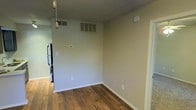 This single-story space is finished with warm-toned hardwood-style flooring that runs throughout, providing a cohesive and inviting foundation. The walls are painted in a neutral, light beige tone, creating a clean and versatile backdrop adaptable to various décor styles. The textured popcorn ceiling adds a classic touch, while multiple HVAC vents — including both supply and return air vents — ensure efficient climate control throughout the space. A wall-mounted thermostat offers convenient temperature management. White baseboards neatly frame the perimeter of the room, adding a crisp, finished look. A pull-chain light fixture is visible, offering straightforward overhead lighting. The layout opens toward an adjacent area where a stainless steel refrigerator is partially visible, indicating a connected kitchen space. Electrical outlets are positioned at standard intervals along the walls, offering practical functionality. The overall ambiance is clean, neutral, and move-in ready, providing a blank canvas well-suited for a variety of uses and personal styles.