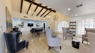 This single-floor space is defined by its striking architectural character and thoughtfully curated interior design. The main living area commands immediate attention with its vaulted ceiling adorned with bold, dark wood exposed beams that run the length of the room, creating a dramatic sense of height and volume. Dual orb-style chandeliers provide warm, ambient lighting, while a large decorative clock mounted on the crisp white walls adds a bold focal point. Arched multi-pane windows allow natural light to filter in and offer views of the outdoors, enhancing the open, airy atmosphere throughout.

Light wood-look flooring runs continuously across the space, complementing the neutral white walls and providing a clean, cohesive foundation. The main area is furnished with wingback chairs, a tufted leather sofa, and a sturdy industrial-style trestle table with bar stools, creating versatile zones for lounging and gathering. A wall-mounted television and built-in fireplace with white subway tile surround add functional and aesthetic appeal to a secondary seating area.

An adjacent room showcases a series of four tall vertical abstract paintings, contributing an artistic touch. The kitchen features dark cabinetry, light granite countertops, and stainless steel appliances, offering a practical and well-appointed cooking space.