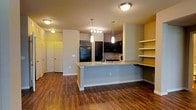 This single-floor residence showcases a thoughtfully designed open-concept layout that seamlessly connects the kitchen and living areas. The kitchen is a standout feature, centered around a large peninsula with granite countertops that offers generous prep space and casual seating. Dark cabinetry lines the kitchen walls, providing ample storage and lending a sleek, modern aesthetic. Stainless steel and black appliances, including a full-size refrigerator, complement the contemporary design. Pendant lighting hangs above the peninsula, while flush-mount ceiling fixtures illuminate the broader living space, creating a warm and welcoming ambiance. Rich hardwood-style flooring extends throughout the main living area, adding warmth and visual continuity to the space. Open display shelving near the living area provides both functional storage and an opportunity for personalized décor. A built-in desk area with matching countertop surface adds a practical workspace element to the layout. The neutral wall tones, accented by soft yellow hues, contribute to a bright and inviting atmosphere. The interior flows efficiently from the entry to the main living space, making this residence as functional as it is visually appealing.