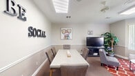 Located at 1900 Bella Vista Way in Port St. Lucie, Florida, this inviting social room is thoughtfully designed for gathering, relaxing, and connecting. The space is divided into two functional zones that work harmoniously together. On one side, a white rectangular table surrounded by upholstered dining chairs provides an ideal setting for games, meetings, or casual meals, accented by bold "BE SOCIAL" wall lettering that sets the tone for the room. On the other side, a comfortable lounge area features tufted gray sofas and accent chairs arranged around a round marble-top coffee table, all anchored by a vibrant red-and-white patterned area rug. A flat-screen TV mounted on a media console serves both zones, accompanied by a "Relax and Unwind" sign that reinforces the room's welcoming atmosphere. A prominent skylight bathes the interior in natural light, while a sweeping bay window along one wall frames lush greenery views and fills the lounge area with an airy brightness. Warm-toned carpeting, a two-tone wall treatment with chair rail molding, and potted tropical plants throughout add warmth and a touch of nature to the overall ambiance. Hexagonal wall shelves near the entry add a decorative finishing touch to this well-appointed communal space.