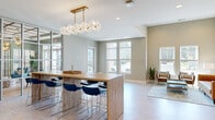 Located at 111 Teal Cir in Little River, South Carolina, this well-appointed community lounge offers a thoughtfully designed, single-floor open-plan layout that seamlessly blends social and functional spaces. The room is anchored by a striking wood-topped bar-height communal table surrounded by modern navy blue chairs, suspended beneath an eye-catching brass-framed globe pendant chandelier that serves as the centerpiece of the space. A full-height steel-framed glass partition wall on one side adds an architectural element while allowing light to pass freely, creating a sense of openness and connectivity. The patterned herringbone-style flooring adds texture and visual depth throughout. A dedicated lounge area features soaring ceilings, a channeled caramel leather sofa, coordinating accent chairs, and a sleek coffee table, all set against neutral warm-toned walls adorned with framed art. Large windows along multiple walls flood the interior with abundant natural light and offer views to the outdoors. An additional conference-style dining table with white chairs and a built-in desk area provide versatile workspace options. Recessed lighting complements the designer pendant fixture, ensuring the space is well-lit throughout the day and evening.