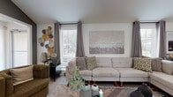 This single-floor community amenity space is thoughtfully designed to offer residents a comfortable and functional environment for both relaxation and productivity. The lounge area serves as the heart of the space, featuring a generously sized L-shaped sectional sofa with mid-century modern legs, complemented by a pair of olive-green barrel accent chairs arranged around a glass-top side table. Natural light pours in through large windows dressed with gray drapes, while decorative wall art and a collection of woven wall discs add texture and warmth to the neutral color palette. Adjacent to the lounge, a kitchenette station is outfitted with white cabinetry, stainless steel appliances, a double sink, and a Starbucks-branded coffee machine — providing a convenient refreshment area for residents. A wall-mounted television above the counter adds to the communal atmosphere. The fitness center is visible through floor-to-ceiling glass panels and features a variety of cardio and strength-training equipment, including treadmills and stationary bikes, along with mirrored walls and an overhead skylight that fills the space with additional light. Nearby, a co-working nook provides a quiet setting with a sleek desk and rolling chairs, ideal for focused work or informal meetings. Tile flooring runs throughout the common areas, lending a clean, polished finish to the overall design.