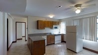 This single-story space centers around a practical and well-equipped kitchen that serves as its standout feature. The kitchen is designed with warm medium-toned wood cabinetry offering generous upper and lower storage, complemented by dark granite-look countertops that add a touch of contrast and durability. A peninsula extends from the main counter, providing additional prep space or casual seating potential. Light-toned ceramic tile flooring runs throughout, brightening the area and offering easy maintenance. White appliances, including a refrigerator, electric range, built-in microwave, and dishwasher, are all included, making the kitchen fully functional. A stainless steel sink sits beneath a window that invites natural light into the cooking area, while additional windows along the adjacent wall further enhance the brightness of the space. A ceiling fan with a light fixture contributes to air circulation and overhead lighting. A doorway visible from the kitchen hints at additional rooms beyond, suggesting a connected, flowing layout throughout the single-floor plan. The overall ambiance is clean, practical, and move-in ready.