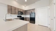 This single-floor residence presents a clean, contemporary design highlighted by light-toned tile flooring that flows consistently throughout the entire space. The kitchen serves as a central feature, showcasing medium-dark wood cabinetry extending to elevated heights, a crisp white peninsula with an integrated sink, and a full suite of black appliances including a refrigerator, range, and over-the-range microwave. Recessed lighting illuminates the open-concept kitchen and living area, which benefits from generous ceiling height that contributes to an airy atmosphere. The entryway offers a practical transition point with panel-style interior doors providing access to additional rooms and storage. The bedroom features neutral beige walls, a closet with built-in rod shelving, and a window that invites natural light and outdoor views. A secondary room with its own window maintains the same bright, neutral palette. The bathroom is outfitted with a tiled tub/shower combination, a dark wood vanity with a white countertop, and matching tile flooring, maintaining visual cohesion with the rest of the unit. Overall, the space presents a well-organized, modern aesthetic defined by neutral tones and consistent finishes.