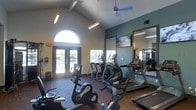 This single-floor fitness center is thoughtfully designed to provide an energizing and functional workout environment. The space is anchored by soaring vaulted ceilings with exposed beams, which lend an open, airy feel and contribute to a sense of generous vertical space. Natural light floods the room through a striking arched window and a set of glass-paneled double doors at the entrance, creating a bright and welcoming atmosphere. Overhead, a ceiling fan and hanging linear light fixtures ensure comfortable airflow and consistent illumination throughout the day. The fitness area is outfitted with a comprehensive selection of cardio equipment, including stationary bikes, elliptical trainers, and treadmills, all arranged to maximize space and ease of use. A large wall-mounted mirror on one side of the room expands the sense of space and allows exercisers to monitor their form, while a flat-screen television is mounted nearby for entertainment during workouts. The flooring features a patterned carpet that adds visual warmth to the space. The teal accent wall provides a modern, energizing color contrast against the neutral tones of the surrounding walls, completing the clean and motivating aesthetic of this well-appointed fitness facility.