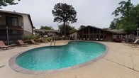 This outdoor amenity area centers around a freeform swimming pool with a distinctly curved, kidney-inspired shape and vivid turquoise water. The pool is framed by a wide, brick-edged concrete deck that provides ample space for circulation and relaxation. Multiple lounge chairs and reclining seats are arranged along the poolside, offering residents comfortable spots to unwind in an open-air setting.

Adjacent to the pool is a single-story clubhouse structure featuring a striking A-frame roofline and large floor-to-ceiling glass panels that allow abundant natural light into the interior. The building's dark wood exterior cladding gives it a warm, lodge-inspired aesthetic that complements the surrounding landscape. Entry to the clubhouse appears accessible directly from the pool deck, reinforcing a seamless indoor-outdoor connection.

The broader setting is defined by mature trees, including a prominent tall pine that provides a natural focal point above the pool area. A wrought iron perimeter fence encloses the space, ensuring privacy and security. The landscaping incorporates greenery and shrubs that soften the hardscape and add to the overall welcoming ambiance of this community outdoor amenity space.