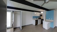 This single-floor space showcases a distinctive mid-century modern aesthetic, defined by its striking exposed black ceiling beams set against white tongue-and-groove paneling. The open layout creates a seamless flow between living and functional areas, offering a clean and airy atmosphere throughout.

The kitchen is thoughtfully designed with crisp white cabinetry, a vibrant turquoise tile backsplash that adds a pop of color, and stainless steel appliances including a dishwasher, electric range, and over-the-range microwave. The countertops and sink are neatly integrated into the layout, and pendant lighting overhead adds both function and visual warmth to the space.

The flooring throughout is a light-toned wood laminate or hardwood, contributing to the bright and cohesive feel of the interior. A built-in closet with a hanging rod provides convenient storage and is positioned to maximize the usability of the open floor plan.

The combination of bold architectural ceiling details, modern finishes, and a carefully curated color palette gives this space a unique character that balances vintage-inspired design with contemporary functionality. The overall ambiance is clean, minimalist, and inviting — ideal for those who appreciate thoughtful design details.