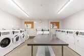 Located at 74 Curlew Ave in Toronto, Ontario, this shared laundry facility is a well-organized, functional space designed to serve multiple users efficiently. The room is finished with light-toned tile flooring and crisp white walls, creating a clean and bright environment enhanced by overhead fluorescent lighting that illuminates the entire space evenly.

The facility is equipped with multiple front-loading washing machines, a top-loading washer, and a row of front-loading dryers, all finished in white and fitted with digital card-payment interfaces for convenient, cashless operation. A utility sink station is also present, offering additional washing options. At the center of the room, a large stainless steel folding and sorting table provides ample workspace for handling laundry.

Two cork bulletin boards mounted on the walls display notices and information for users, while a wall clock keeps track of time. A waste bin and a dedicated coin/card machine are conveniently accessible near the entrance. The space also features a wall-mounted HVAC unit to maintain comfortable temperatures year-round. The single entry door is sturdy and functional, ensuring secure access. Overall, this laundry facility is thoughtfully laid out to maximize usability, cleanliness, and convenience for its users.