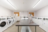 Located at 74 Curlew Ave in Toronto, Ontario, this shared laundry facility is a well-organized, functional space designed to serve multiple users efficiently. The room is finished with light-toned tile flooring and crisp white walls, creating a clean and bright environment enhanced by overhead fluorescent lighting that illuminates the entire space evenly.

The facility is equipped with multiple front-loading washing machines, a top-loading washer, and a row of front-loading dryers, all finished in white and fitted with digital card-payment interfaces for convenient, cashless operation. A utility sink station is also present, offering additional washing options. At the center of the room, a large stainless steel folding and sorting table provides ample workspace for handling laundry.

Two cork bulletin boards mounted on the walls display notices and information for users, while a wall clock keeps track of time. A waste bin and a dedicated coin/card machine are conveniently accessible near the entrance. The space also features a wall-mounted HVAC unit to maintain comfortable temperatures year-round. The single entry door is sturdy and functional, ensuring secure access. Overall, this laundry facility is thoughtfully laid out to maximize usability, cleanliness, and convenience for its users.