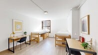 Located at 97 Columbia Heights in the Brooklyn Heights neighborhood of Brooklyn, NY, this compact single-floor unit offers a thoughtfully organized layout across a living room, bedroom, and full bathroom. The interiors are defined by a clean, minimalist aesthetic, with crisp white walls, light wood-toned flooring, and track lighting that keep the space feeling bright and cohesive throughout.

The bedroom accommodates two twin beds with built-in drawer storage beneath, maximizing utility within the cozy footprint. A wood-framed window invites natural light and provides views of the surrounding Brooklyn streetscape, while a baseboard heating unit ensures year-round comfort. Each sleeping area is paired with a dedicated desk and task lighting, making the room well-suited for both rest and focused work.

The living area extends the functional layout, featuring an open workspace with a desk and chair alongside a wall-mounted clothing rack that serves as an open wardrobe solution. Trailing greenery adds a touch of natural warmth to the otherwise clean, modern palette.

The bathroom is finished in classic white subway and mosaic tile, featuring a full soaking tub with shower, a pedestal sink with chrome fixtures, and a large wall mirror. The all-white finish reinforces the unit's consistent, streamlined aesthetic from room to room.