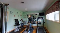 Located at 1061 Johnnie Dodds Blvd in Mt. Pleasant, South Carolina, this single-floor fitness center offers a well-equipped and inviting space for exercise and wellness. The room features a curated selection of cardio machines, including a treadmill, elliptical trainer, and stationary bike, all arranged to maximize usability and comfort. A large wall-mounted mirror spans one side of the room, creating a sense of openness and allowing users to monitor their form during workouts. A flat-screen television is mounted on the wall, providing entertainment during exercise sessions. The space is finished with warm hardwood-style flooring and recessed lighting in the white ceiling, contributing to a clean and energetic ambiance. The walls are painted in a soothing sage green and are decorated with playful, colorful decals and motivational wall art, adding a fun and encouraging personality to the environment. A window allows natural light to filter into the space. The overall design strikes a balance between functional fitness facility and a lively, approachable atmosphere, making it a comfortable destination for daily exercise routines.
