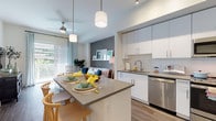 This contemporary single-floor residence is thoughtfully designed with a clean, modern aesthetic throughout. The open-concept kitchen is a standout feature, outfitted with crisp white cabinetry extending to the ceiling, stainless steel appliances including a French door refrigerator, a gas range, microwave, and dishwasher. A large central island with a quartz-style countertop provides additional seating and prep space, illuminated by stylish pendant lighting and complemented by a subway tile backsplash. Wood-look flooring flows seamlessly through the kitchen and into the living area, where a charming accent wall adds depth and character. The living room benefits from abundant natural light through a full-width sliding glass door that opens to an outdoor balcony with greenery views, and a ceiling fan enhances the airy feel. The bedroom is generously proportioned with elevated ceilings, a ceiling fan, and direct access to a walk-in closet, creating a comfortable and functional retreat. The bathroom features a dual-sink vanity with white cabinetry, quartz countertops, a large framed mirror, and a glass-enclosed walk-in shower with white tile surround. Every detail of this residence reflects a consistent, modern design sensibility with quality finishes throughout.
