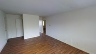 This single-floor space presents a clean, minimalist aesthetic defined by crisp white walls and smooth white ceilings that help maximize the sense of openness throughout. The rich, dark hardwood-style flooring runs continuously across the main living area, providing a warm contrast to the bright, neutral walls and contributing to a welcoming atmosphere.

The main living area is generously proportioned, offering ample room for a variety of furniture arrangements. White baseboards frame the perimeter of the space, adding a polished finishing detail. A doorway visible from the main room leads into an adjacent area, where a window allows natural light to filter through, brightening the interior and providing a visual connection to the outside.

The layout appears designed for practical flow between spaces, with the open sightlines lending the interior a sense of continuity. A light switch and electrical outlet are positioned conveniently along the walls. The overall condition of the space appears well-maintained, with the neutral palette offering a versatile backdrop suitable for a range of interior styles and uses.