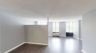 This single-floor unit offers a practical and well-designed layout that balances open living spaces with functional private areas. Hardwood-style flooring flows consistently throughout the main living areas, complemented by neutral gray walls and textured ceilings that contribute to a clean, contemporary aesthetic.

The living area is notably bright, featuring a bank of large windows fitted with horizontal blinds that allow generous natural light to fill the room. A wall-mounted PTAC unit provides climate control, and the open configuration of the space allows for flexible furniture arrangement.

The kitchen is efficiently designed and well-equipped, showcasing rich dark-wood cabinetry, granite-style countertops, a stainless steel refrigerator, built-in microwave, electric range, and a single-basin sink. The tile backsplash adds a refined detail to the cooking area, which opens directly toward the main living and dining space.

A hallway connects the living areas to the private spaces, including closeted storage and a full bathroom. The bathroom features a dark-wood vanity with a granite-topped sink, a mirrored medicine cabinet, tile-surround tub and shower combination, and hardwood-style flooring that mirrors the rest of the unit, creating a cohesive feel throughout.