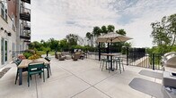 Located at 7689 Hardwood Ave S in Cottage Grove, Minnesota, this generously sized outdoor amenity area offers a well-appointed community space designed for relaxation and social gathering. The space is anchored by a raised rectangular pool with stone-clad exterior walls, accessible via stainless steel handrails and surrounded by a row of chaise lounge chairs for sunbathing. The expansive concrete patio extends across the grounds, providing ample open space for movement and activity. A dedicated lounge area features wicker seating arranged around a central fire table, creating an inviting setting for conversation and relaxation. High-top bistro tables with stools and a shaded umbrella offer an additional gathering spot suited for casual outdoor dining or socializing. A separate dining area adjacent to the building includes a full-sized outdoor table with seating for six, positioned near large glass entry doors and exterior wall-mounted lighting. The entire space is enclosed by sleek black iron fencing, lending a clean, modern aesthetic while maintaining an open feel. Mature trees line the perimeter beyond the fencing, providing a natural backdrop and a sense of privacy. The contemporary multi-story building facade, featuring a combination of stone, panel siding, and warm terracotta tones, frames the space and reinforces the modern residential character of the community.