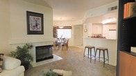 This single-story residence showcases an open-concept design that seamlessly connects the living, dining, and kitchen areas into one cohesive, flowing space. The living room serves as the focal point, anchored by a tiled fireplace with a sleek black surround, complemented by neutral-toned carpet and warm, cream-colored walls. A framed artwork piece hangs above the fireplace, adding a decorative touch to the space.

The dining area sits adjacent to the living room, accommodating a full dining table and chairs, and is illuminated by a pendant light fixture. Vertical blinds on the nearby window allow for natural light control while maintaining privacy.

The kitchen features white cabinetry and is accessible via a raised breakfast bar counter, equipped with two bar stools — an ideal spot for casual dining or conversation. The pass-through design of the kitchen enhances the open feel of the overall layout.

Throughout the space, the consistent use of light, neutral tones creates a bright and airy ambiance. The combination of tile near the fireplace and carpet in the living areas adds texture and visual interest to the single-level floor plan.