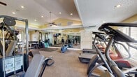 Located in Saint Petersburg, Florida, this fitness center offers a comprehensive workout facility within a single, well-appointed exercise room. The space features vaulted ceilings with a warm yellow accent trim, a textured ceiling, recessed lighting, and ceiling fans that contribute to a bright and comfortable atmosphere. Large windows along the walls allow natural light to fill the room, creating an open and energizing environment.

The exercise room is thoughtfully organized to accommodate a wide range of fitness activities. Cardio equipment includes treadmills, elliptical machines, and a stationary bike, all positioned near the windows to take advantage of the natural light and outdoor views. A dedicated strength training area houses a multi-station cable machine, weight benches, and a full rack of dumbbells, catering to users of varying fitness levels. Open floor space with exercise mats provides ample room for stretching, yoga, or floor-based workouts, along with stability balls and other accessories.

Full-length mirrors line a significant portion of the walls, visually expanding the space and allowing users to monitor their form during exercise. The carpeted flooring adds a layer of comfort underfoot, while the glass-paneled door at the entrance provides easy access. The overall ambiance is clean, functional, and inviting, making this fitness center a practical and well-rounded amenity.