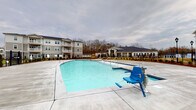 Located at 4739 Starr Ranch Rd in Indian Land, South Carolina, this outdoor amenity space is designed to offer residents a well-appointed recreational environment. At the center of the area is a generously sized, resort-style swimming pool with crystal-clear blue water and a wide surrounding concrete deck that provides ample space for lounging. An ADA-compliant pool lift is installed poolside, ensuring accessibility for all residents.

Adjacent to the pool is a substantial covered pavilion constructed with natural wood timber framing and an exposed wood-plank pitched roof, lending a warm and rustic character to the outdoor setting. The pavilion shelters a large concrete patio area and features a built-in stone outdoor fireplace, crafted with stacked stone masonry, creating a natural gathering focal point. A stone bench is integrated into the fireplace surround, offering additional seating.

The surrounding landscape includes freshly planted evergreen shrubs and young trees, enclosed by a black iron fence that defines the amenity area while maintaining open sightlines to the multi-story residential buildings beyond. Decorative lamp posts are positioned throughout the space, enhancing both the aesthetic and functionality of the area after dark. The overall design balances practicality and comfort, making it an inviting outdoor destination for relaxation and community gatherings.