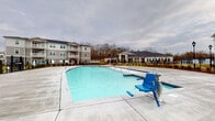 Located at 4739 Starr Ranch Rd in Indian Land, South Carolina, this outdoor amenity space is designed to offer residents a well-appointed recreational environment. At the center of the area is a generously sized, resort-style swimming pool with crystal-clear blue water and a wide surrounding concrete deck that provides ample space for lounging. An ADA-compliant pool lift is installed poolside, ensuring accessibility for all residents.

Adjacent to the pool is a substantial covered pavilion constructed with natural wood timber framing and an exposed wood-plank pitched roof, lending a warm and rustic character to the outdoor setting. The pavilion shelters a large concrete patio area and features a built-in stone outdoor fireplace, crafted with stacked stone masonry, creating a natural gathering focal point. A stone bench is integrated into the fireplace surround, offering additional seating.

The surrounding landscape includes freshly planted evergreen shrubs and young trees, enclosed by a black iron fence that defines the amenity area while maintaining open sightlines to the multi-story residential buildings beyond. Decorative lamp posts are positioned throughout the space, enhancing both the aesthetic and functionality of the area after dark. The overall design balances practicality and comfort, making it an inviting outdoor destination for relaxation and community gatherings.