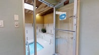 This single-level indoor aquatic facility offers a self-contained and functional recreational environment. The space is accessed through a set of glass-paneled entry doors, providing a clear view of the interior before entry. Upon entering, visitors are greeted by a compact pool or hot tub with clean, blue water, set within a tiled surround featuring decorative mosaic accent strips in contrasting colors. The walls are finished with white tile accented by vertical bands of colored mosaic tile, lending the space a clean and orderly aesthetic. A built-in ledge or bench area lines the perimeter, offering a convenient place to sit or set belongings poolside. A potted plant adds a subtle natural touch to the otherwise utilitarian environment. The ceiling features a warm-toned wood or paneled finish, complemented by skylights or translucent panels that allow natural light to filter into the space. Signage within the space indicates standard facility rules, including a prohibition on glass and alcohol. A dressing room is accessible directly from the pool area, adding to the convenience and functionality of the facility. The overall ambiance is clean, compact, and purpose-built for relaxation and aquatic use.