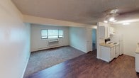 This single-floor residential unit offers a practical and well-organized layout designed for comfortable everyday living. The open living area serves as the heart of the home, combining a carpeted lounge zone with rich dark hardwood-style flooring extending toward the kitchen. A wide window allows natural light to fill the space, and a baseboard heating unit along with a wall-mounted AC unit ensures year-round climate control. A ceiling fan adds additional ventilation to the living area.

The kitchen is positioned adjacent to the living space and features white cabinetry, a countertop with sink, and efficient use of the available area. The transition between rooms is smooth and open, creating a connected flow throughout the unit.

The bedroom is a well-proportioned, carpeted room with neutral walls and a textured ceiling. A window fitted with horizontal blinds provides outdoor views and natural light, while a baseboard heater runs along the exterior wall. The bedroom also benefits from a generously sized walk-in closet equipped with dual hanging rods and a built-in shelving unit, offering ample storage and organization options.

The bathroom includes a full-size shower/tub combo with white surround, a toilet, and a vanity sink, all finished in clean, light tones. Throughout the unit, the textured ceilings, neutral color palette, and practical finishes create a clean, functional living environment.