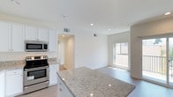 This single-floor unit presents a clean, contemporary design characterized by bright white walls, recessed lighting, and a cohesive neutral palette throughout. The kitchen serves as a functional centerpiece, featuring white shaker-style cabinetry extending to the ceiling, granite countertops, and a full suite of stainless steel appliances including a refrigerator, electric range, built-in microwave, and dishwasher. A kitchen island with matching granite provides additional prep and counter space, while light wood-tone flooring ties the kitchen seamlessly into the adjacent living area. The living space is open and airy, with a sliding glass door leading to a private balcony, allowing for natural light and outdoor views. Bedrooms are carpeted in a dark grey tone and feature large windows that bring in natural light, with views of surrounding brick architecture. Two full bathrooms maintain the unit's consistent aesthetic, each offering white vanity cabinetry, granite countertops, large mirrors, and a shower-tub combination enclosed by sliding glass doors. Tile flooring and a high clerestory window in one bathroom add practical and architectural detail. Overall, the space presents a move-in-ready, modern environment with quality finishes throughout.