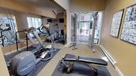 Located at 4700 176th St SW in Lynwood, Washington, this fitness center is a well-equipped, single-floor exercise facility designed to support a comprehensive workout experience. The space features vaulted ceilings with exposed wood beam detailing and skylight panels that allow natural light to brighten the interior, creating an open and energizing atmosphere.

The cardio zone is outfitted with multiple elliptical trainers, treadmills, and a recumbent stationary bike, all positioned alongside a full-wall mirror that visually expands the space. Mounted flat-screen televisions above the mirrors provide entertainment during workouts. A multi-function cable resistance machine serves as the centerpiece for strength training, while a padded workout bench, stability balls, and exercise mats offer versatility for a range of fitness routines.

Instructional fitness and yoga posters are displayed on the warm, golden-toned walls, providing guidance for stretching and supplemental exercises. Large glass doors open directly to an outdoor deck, offering views of surrounding greenery and fresh air during workouts. Carpeted flooring with rubberized mat zones throughout the room provides comfort and traction. The space is finished with baseboard heating and recessed lighting, ensuring year-round usability and a comfortable environment for residents.