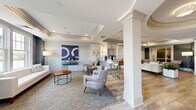 Located at 890 S Matlack St in West Chester, PA, this refined single-floor commercial space makes a striking first impression with a grand rotunda-style entry foyer. The circular ceiling recess is adorned with ornate crown molding and elegant flush-mount light fixtures, immediately establishing a polished, upscale ambiance. Light wood-look flooring extends throughout the open-concept layout, complemented by crisp white columns and detailed millwork that lend a sense of architectural sophistication.

The main open area flows seamlessly between distinct functional zones. A casual lounge area features plush seating, a wall-mounted flat-screen television, and a tray ceiling with recessed lighting — ideal for relaxed gatherings or client meetings. Adjacent to the lounge, a bar-height seating area with sleek modern stools provides an informal workspace or social hub. Bold blue accent artwork punctuates the neutral color palette, adding visual interest throughout.

A dedicated workstation area with a substantial wood executive desk sits beneath another coffered tray ceiling, surrounded by large-scale wall art that reinforces the space's contemporary character. Double blue doors serve as a striking interior focal point, connecting zones while maintaining a sense of grandeur. Large windows along the perimeter allow natural light to fill the space. The overall design balances professionalism with warmth, making this West Chester location a highly functional and visually impressive commercial environment.