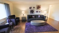 This single-floor space showcases a thoughtfully designed living area that balances contemporary style with warm, inviting elements. The room features rich, dark hardwood flooring that anchors the space and provides a striking contrast against the soft, neutral-toned walls. A tufted charcoal sofa serves as the centerpiece of the seating area, complemented by a matching dark armchair with nailhead trim detailing, adding a refined touch to the overall aesthetic. A large oval silver ottoman rests on a plush, vibrant purple shag area rug, introducing a bold pop of color and texture that draws the eye. Coordinating purple throw pillows on the sofa echo this color scheme throughout the room. Flanking the seating arrangement, elegant table lamps on sleek metallic side tables provide warm ambient lighting. A curated gallery wall of floral artwork in purple and burgundy tones ties the color palette together cohesively. The overall ambiance is stylish and polished, with a deliberate interplay of deep, moody tones and softer neutral backdrops creating a space that feels both comfortable and visually dynamic.